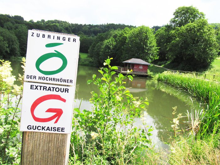 Markierung der Gucksaisee-Tour vor einem Gewässer mit Hütte im Hintegrund